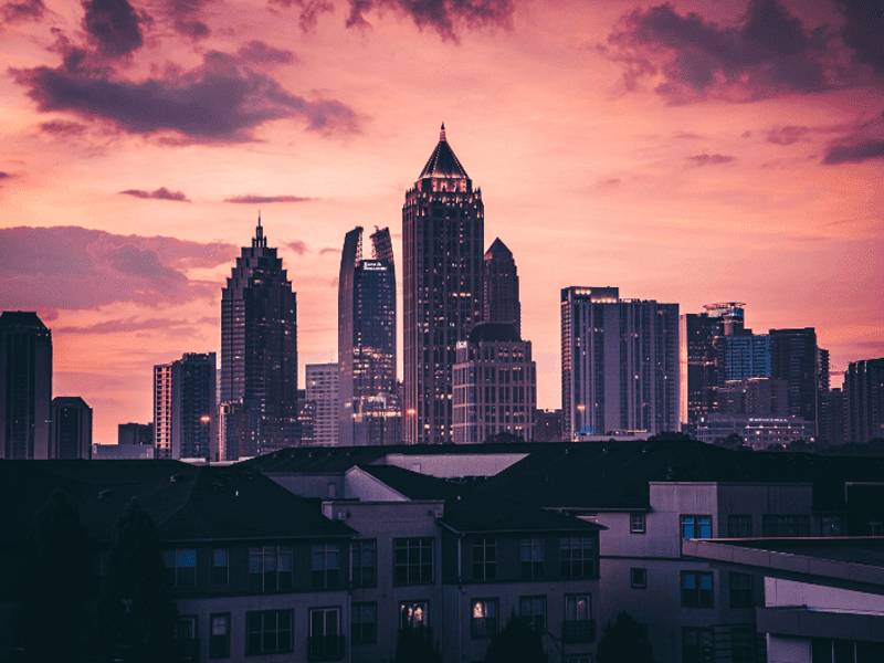 A city skyline with tall skyscrapers at sunset, the sky glowing with shades of pink and purple, and dark apartment buildings in the foreground.