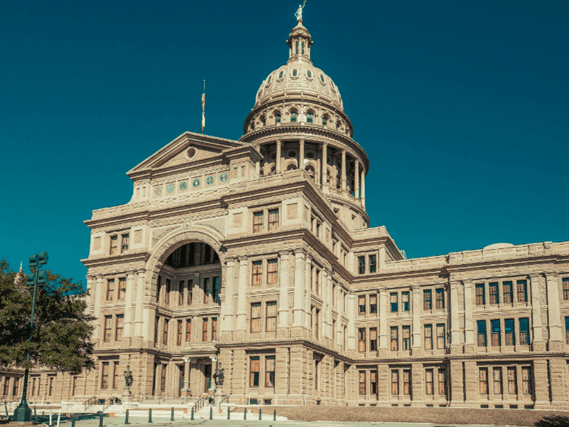 The Texas State Capitol building in Austin, featuring a large central dome, ornate architectural details, and tall arched windows under a clear blue sky.