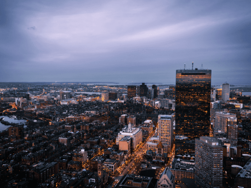 Aerial view of a cityscape at dusk with illuminated buildings and streets. A tall, dark skyscraper with many glowing windows stands prominently, while the sky is overcast with soft purple and blue hues.