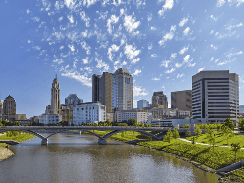 A city skyline with tall office buildings and skyscrapers stands behind a river with a bridge. Green lawns and trees line the riverbanks under a bright blue sky with scattered clouds.
