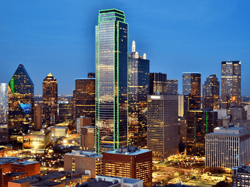 Dallas city skyline at dusk, featuring tall modern skyscrapers with the tallest building outlined in green lights against a blue sky.