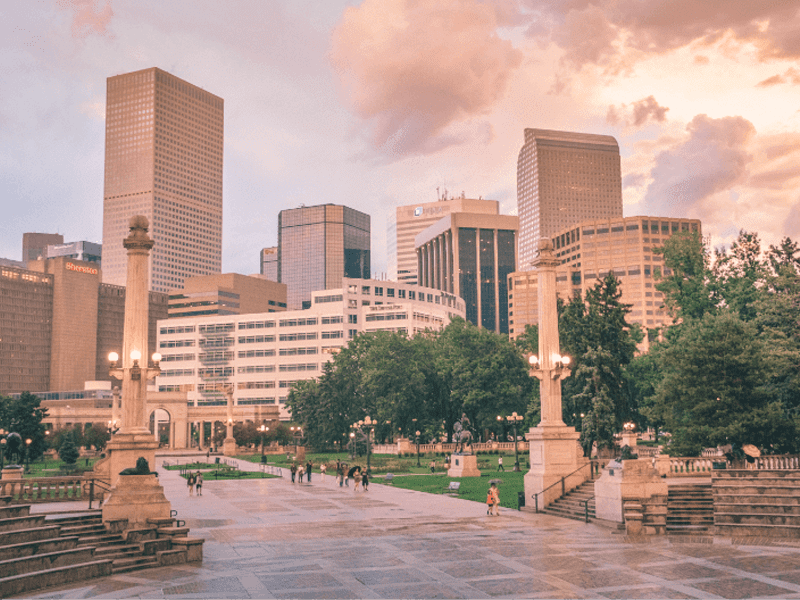 Downtown cityscape with tall modern buildings, a park with trees, people walking, and ornate street lamps at sunset under a cloudy sky.