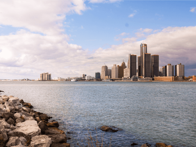 A view of a city skyline with tall buildings along the waterfront, seen across a large river with rocks in the foreground under a partly cloudy sky.