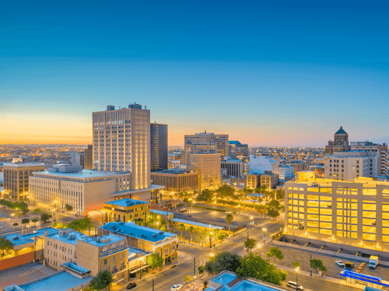 Aerial view of downtown El Paso, Texas at sunset, with tall buildings, illuminated streets, and a clear sky transitioning from blue to orange.
