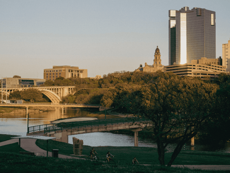 A cityscape with tall office buildings, a historic courthouse, and a river with a curved pedestrian bridge; trees and people biking in the foreground under a clear sky at sunset.