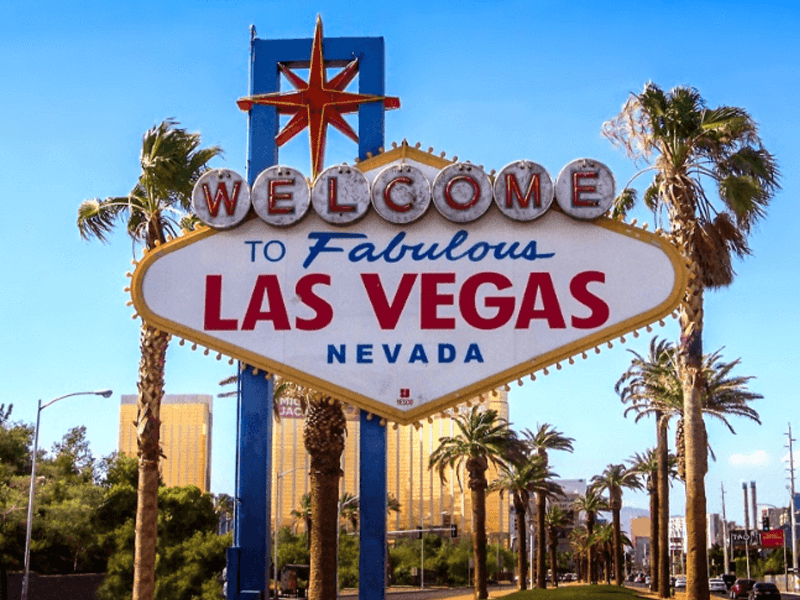 The iconic Welcome to Fabulous Las Vegas, Nevada sign stands tall against a clear blue sky, surrounded by palm trees and with city buildings visible in the background.