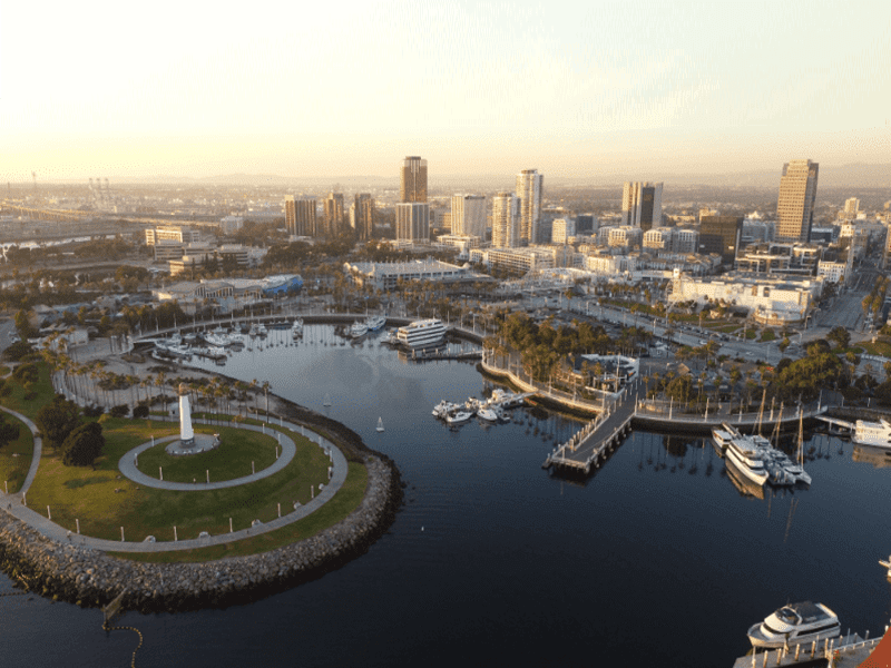 Aerial view of a marina with boats docked, a circular park with a lighthouse, and a city skyline in the background under a hazy sky.