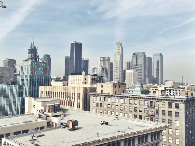 A cityscape of downtown Los Angeles features tall modern skyscrapers, historic buildings, and a blue sky with light clouds. The Eastern Columbia Building stands out with its turquoise art deco design.