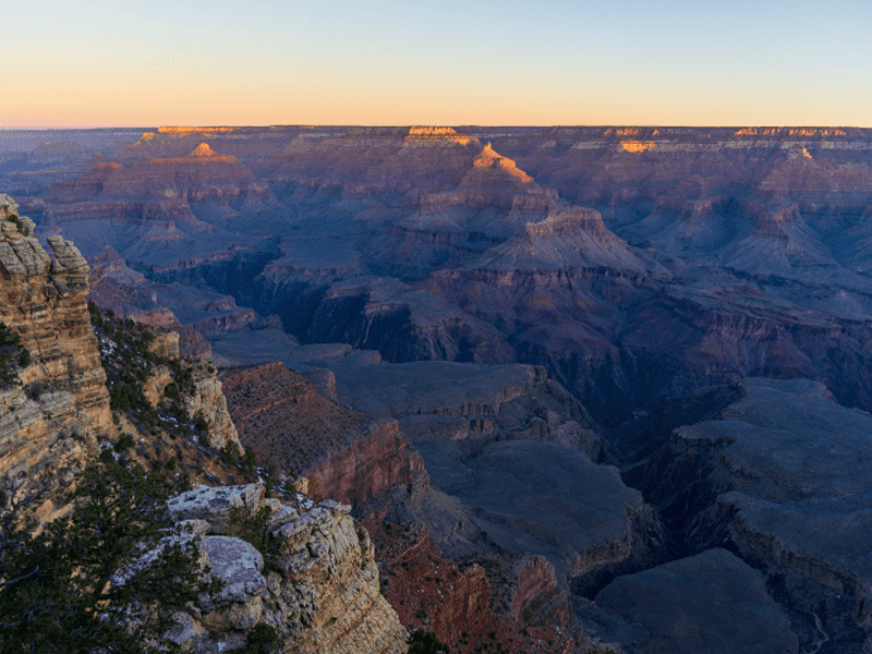 View of the Grand Canyon at sunset, with layered rock formations and deep canyons bathed in warm golden light. The sky is clear, fading from blue to a soft orange near the horizon.