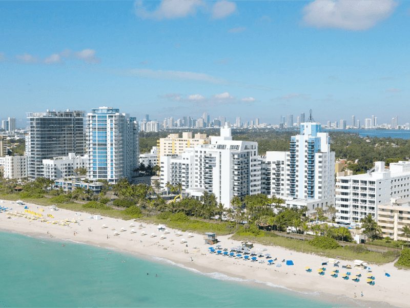 Aerial view of a sandy beach lined with palm trees and colorful umbrellas, with high-rise hotels and buildings behind it. A city skyline is visible in the background under a blue sky with scattered clouds.