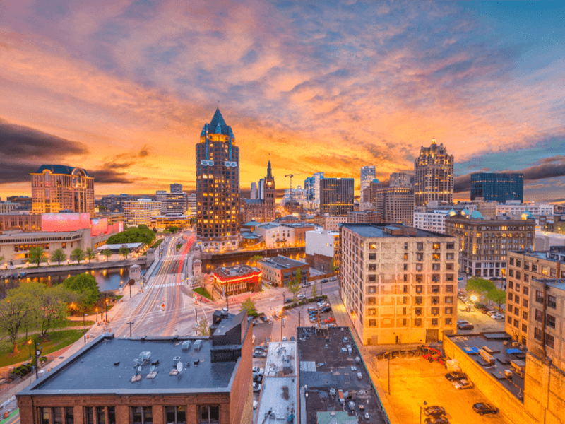 A vibrant cityscape of Milwaukee, Wisconsin at sunset, featuring tall buildings, illuminated streets, and a colorful sky with orange and blue hues. The scene includes the Milwaukee River and various urban structures.