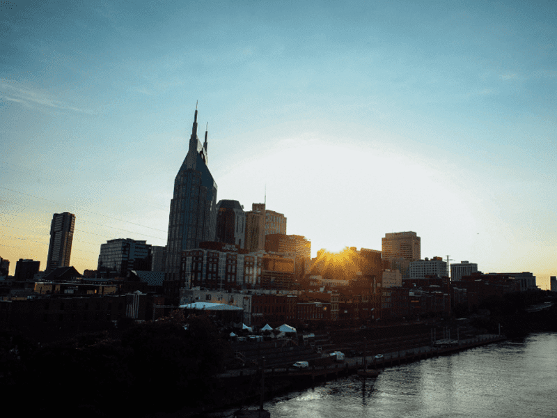 Sunset over a city skyline with tall buildings, including one with a pointed, twin-spire top, and a river in the foreground. The sun is partially hidden behind the buildings.
