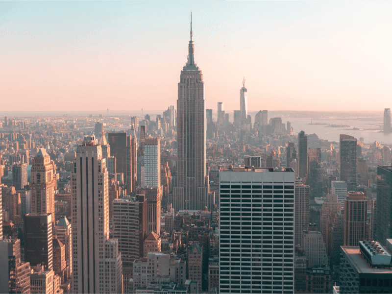 A cityscape view of New York City with the Empire State Building prominently in the center, surrounded by tall skyscrapers under a hazy sky during sunset.