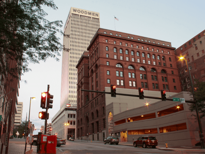 A city street scene with a tall white skyscraper labeled WOODMEN behind a historic red-brick building, traffic lights, parked cars, and a multi-level parking garage in the foreground.