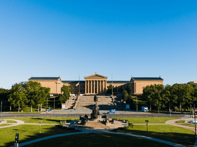 A wide view of the Philadelphia Museum of Art, showing its grand steps, classical columns, and a statue in the foreground, under a clear blue sky.