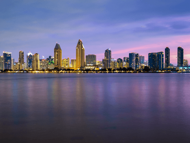 The skyline of a city at dusk with tall buildings illuminated by lights, reflecting on the calm water in the foreground under a purple and blue sky.