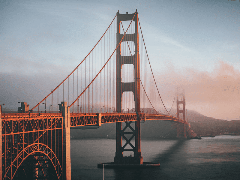 The Golden Gate Bridge in San Francisco spans across foggy blue water, with its tall orange towers rising against a partly cloudy sky and distant hills in the background.