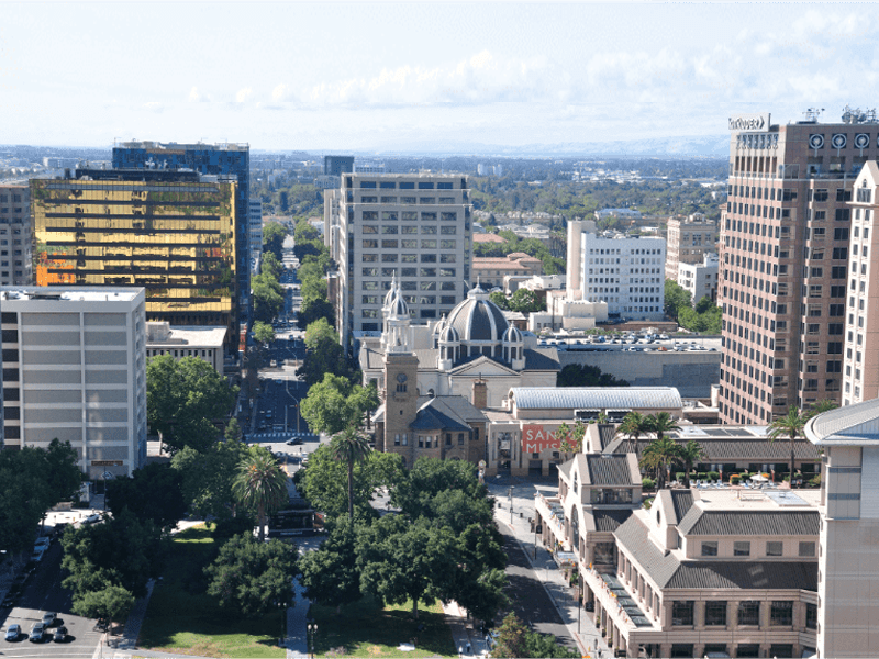 Aerial view of downtown San Jose, California, featuring tall office buildings, a church with a dome, tree-lined streets, and a clear sky in the background.