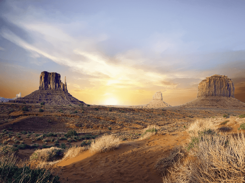 Desert landscape at sunset with sparse shrubs and three large, distinct rock formations rising from the reddish-brown earth under a partly cloudy sky.