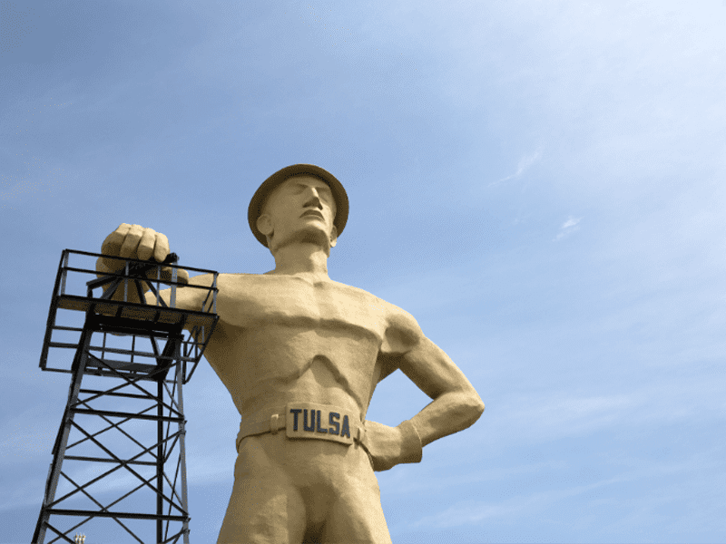 A large statue of a man wearing a hard hat, labeled “TULSA” on his belt, stands with one hand on his hip and the other resting on an oil derrick against a blue sky.