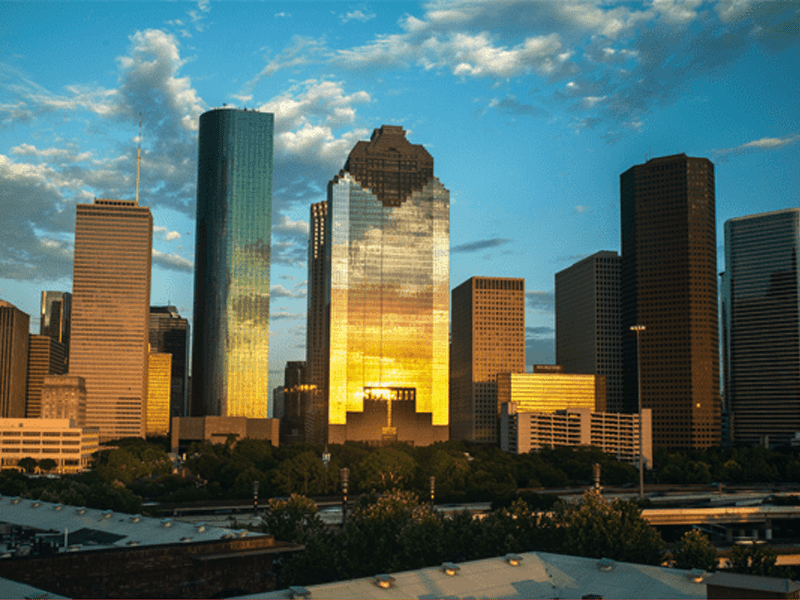 A city skyline at sunset with tall glass skyscrapers reflecting golden sunlight, partly cloudy blue sky above, and trees and buildings in the foreground.