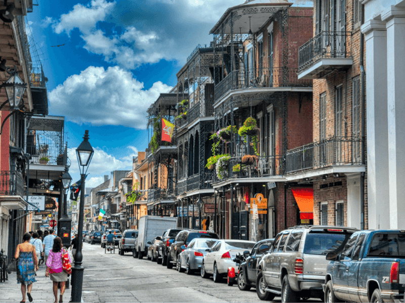 A vibrant city street lined with parked cars, historic buildings with wrought-iron balconies, and greenery. People walk along the sidewalk under a bright blue sky with scattered clouds.