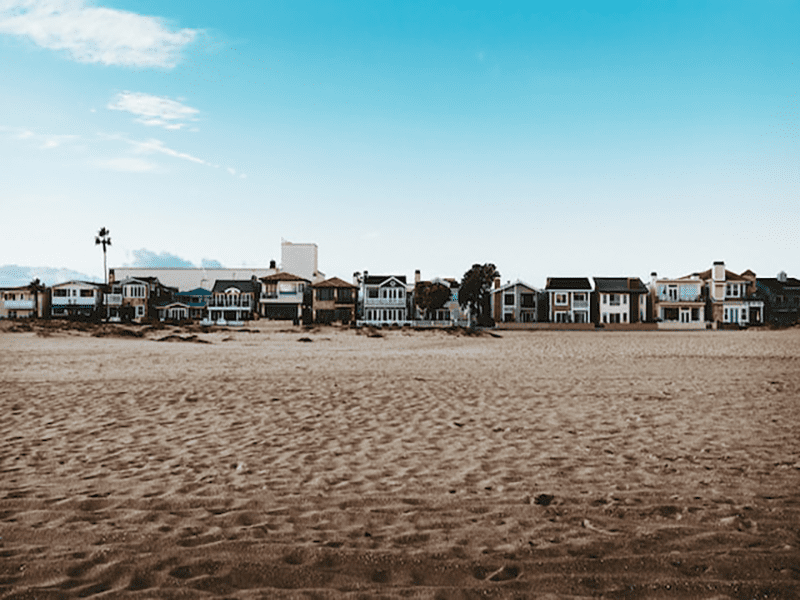 Row of beachfront houses facing a wide, sandy beach under a bright blue sky with scattered clouds. No people are visible, and the scene looks calm and peaceful.