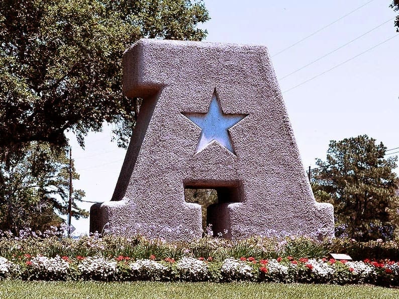 Large stone sculpture of the letter A with a cutout star in the center, surrounded by flowers and grass, with trees in the background.