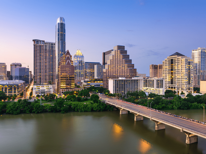 Downtown Austin, Texas skyline at dusk, with tall buildings and skyscrapers lit up, trees along the riverbank, and a bridge crossing the calm river in the foreground.