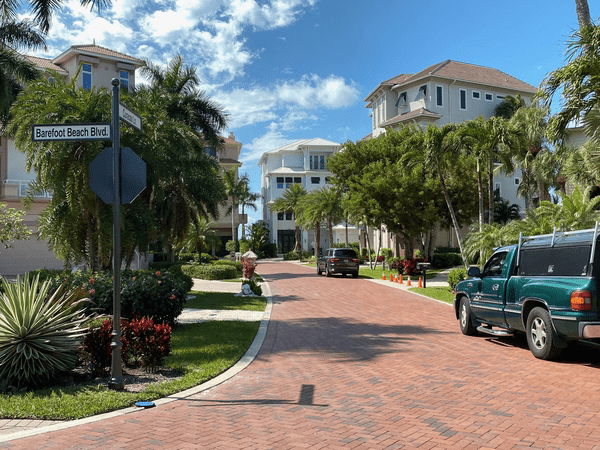 A brick-paved street lined with palm trees and upscale houses under a bright blue sky. Two vehicles are parked on the right, and a street sign reads “Barefoot Beach Blvd.”.