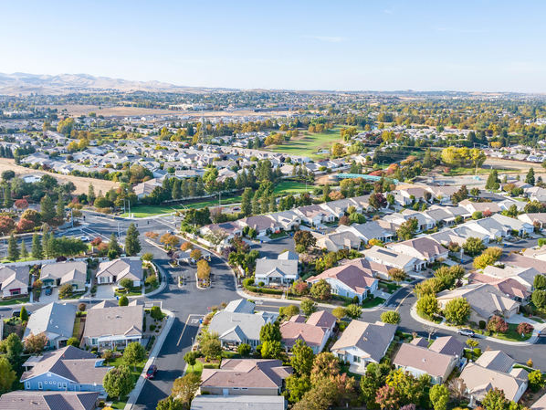 Aerial view of a suburban neighborhood with neatly arranged houses, green lawns, and tree-lined streets, surrounded by open fields and hills in the background under a clear blue sky.