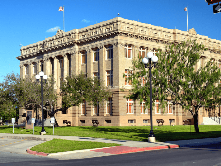 A historic, beige stone courthouse with columns, large windows, and American flags on the roof, surrounded by green lawns, trees, and lampposts, under a clear blue sky.