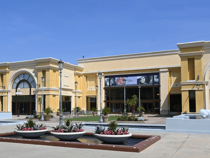 A modern yellow building with arched windows and glass doors stands in bright sunlight. In front, there are planters with flowers, a fountain, and tall street lamps. A movie poster banner hangs above the entrance.