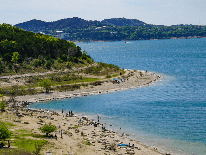 A curved lakeshore with scattered rocks and people walking, relaxing, and playing by the water. Green hills with trees rise in the background under a clear sky.