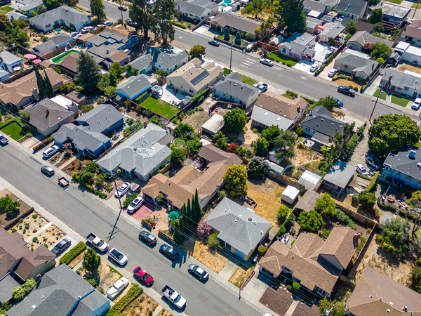 Aerial view of a suburban neighborhood with rows of houses, green lawns, cars parked along the streets, and trees scattered throughout the residential area on a sunny day.
