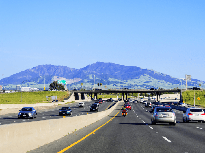 Cars and motorcycles travel on a multi-lane freeway with an overpass in the distance; green hills and a large mountain range appear under a clear blue sky in the background.