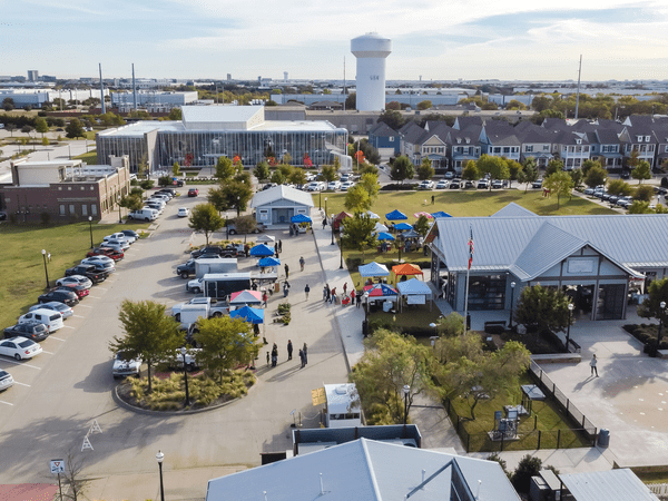 Aerial view of a community event with tents, booths, and people gathered in a parking lot, surrounded by buildings, trees, and a prominent white water tower in the background.