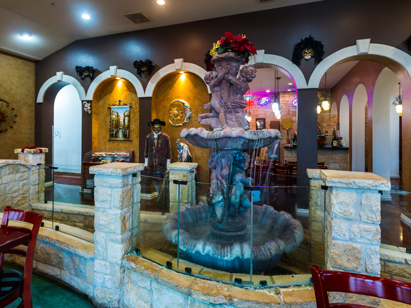 A decorative indoor fountain with cherub statues is surrounded by stone pillars and glass panels in a restaurant with arched walls, warm lighting, and a man in a hat standing in the background.