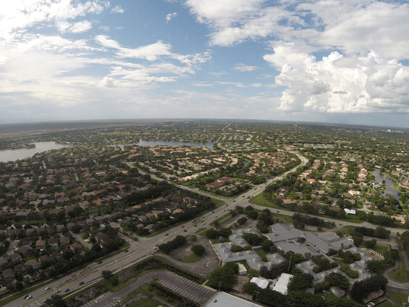 Aerial view of a suburban neighborhood with houses, roads, and patches of greenery under a partly cloudy sky. Lakes and more houses stretch into the distance.