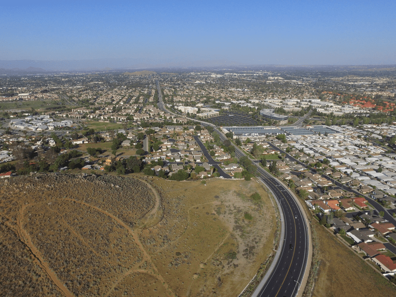Aerial view of a suburban area with houses, roads, and some commercial buildings, bordered by a dry grassy hill in the foreground and a clear blue sky overhead.