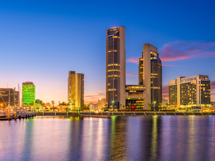 Downtown Corpus Christi, Texas skyline at dusk, with tall buildings lit up, a calm waterfront in the foreground, and a colorful sky with hues of blue, purple, and orange in the background.