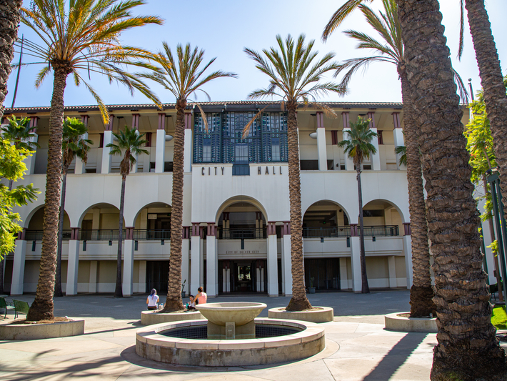 A city hall building with arched entrances, surrounded by palm trees. A round fountain is in the foreground, and a few people are sitting on benches nearby under a sunny sky.