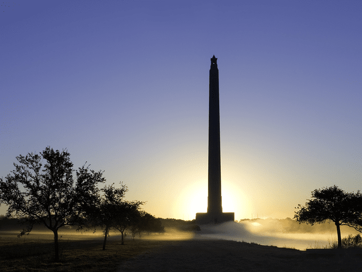 A tall monument is silhouetted against a rising sun, with mist and trees in the foreground, under a clear sky.