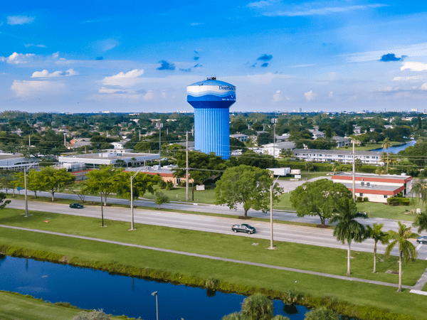 A large blue water tower labeled Deerfield Beach rises above a suburban area with houses, trees, and roads. Several cars drive by, and a canal runs alongside the street under a partly cloudy sky.
