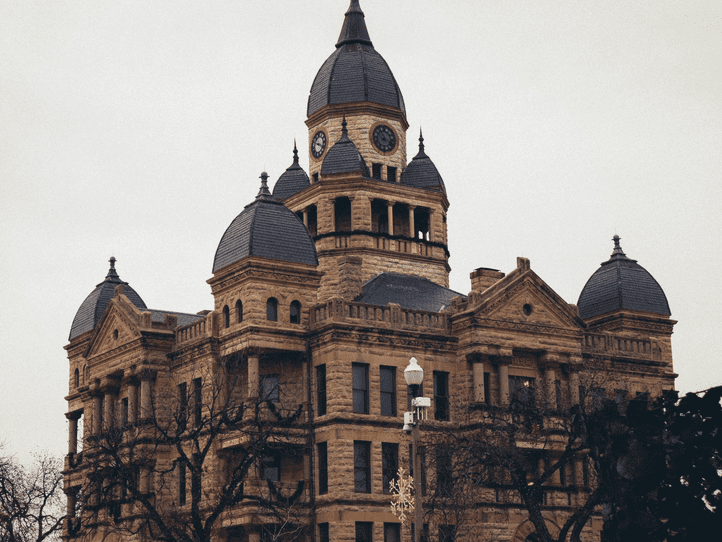 A historic stone courthouse with multiple domed towers and a central clock tower, set against an overcast sky and partially framed by bare trees.