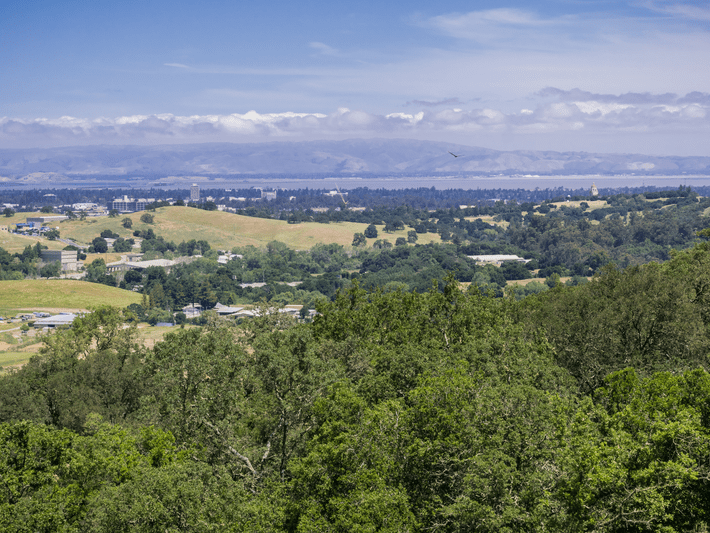 A scenic landscape with green trees and rolling hills in the foreground, scattered buildings, and a distant city. Mountains and a cloudy blue sky appear in the background.