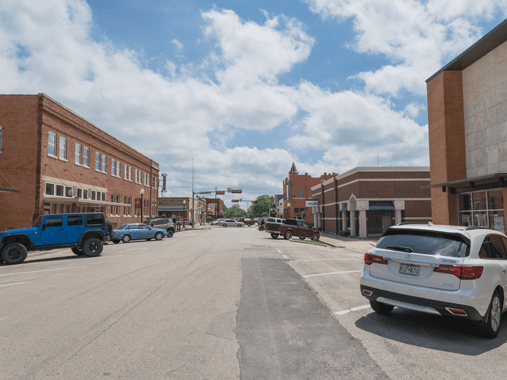 A small town street with parked cars on both sides, red brick buildings, and a partly cloudy sky overhead. The street appears quiet and empty, with no people visible.