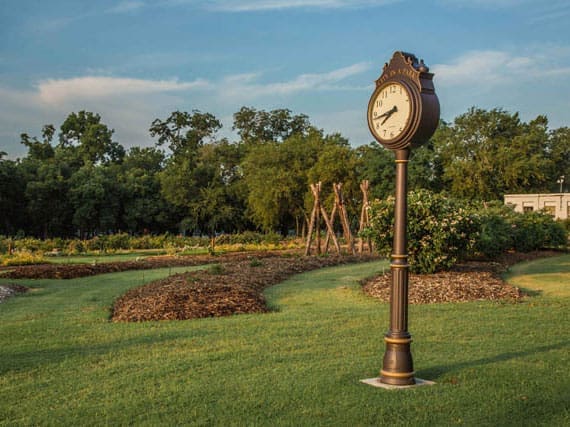 A vintage-style street clock stands on a well-kept lawn in a garden with landscaped flower beds, trees in the background, and a partly cloudy sky overhead.