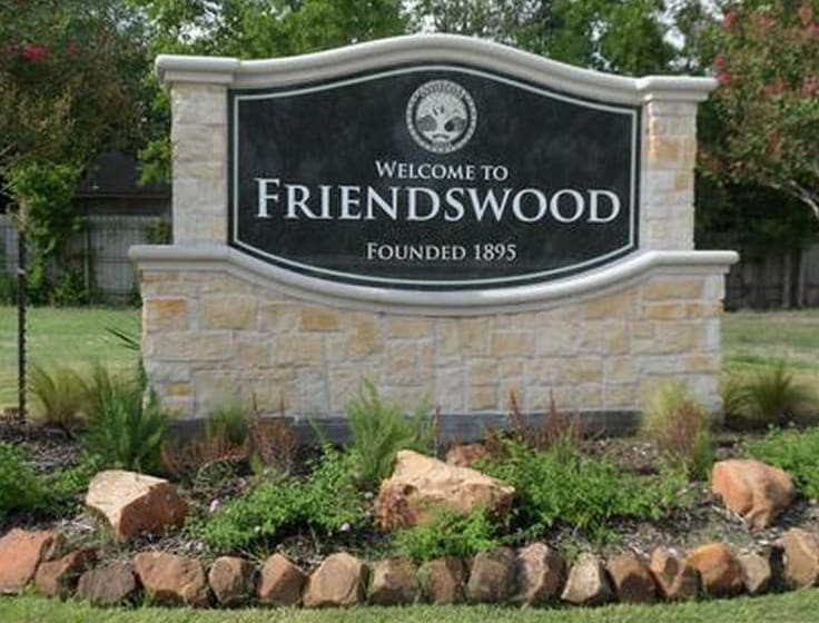 A stone and brick sign reads Welcome to Friendswood, Founded 1895, surrounded by landscaping with rocks, plants, and flowers, with trees and grass in the background.