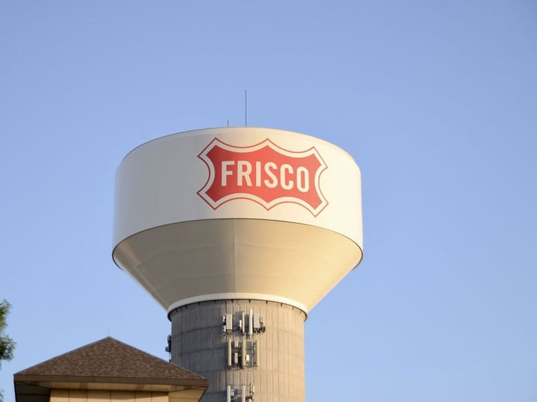 A tall water tower with a white tank displaying a red FRISCO sign stands against a clear blue sky. Part of a building roof and some tree branches are visible at the bottom left.